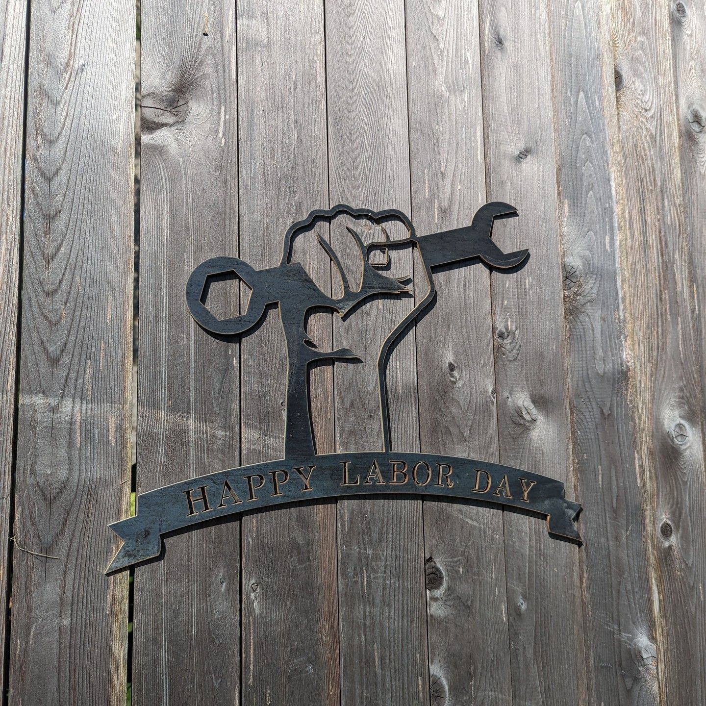 Black metal emblem of a raised fist holding a wrench on weathered wood, with a banner reading “Happy Labor Day.”