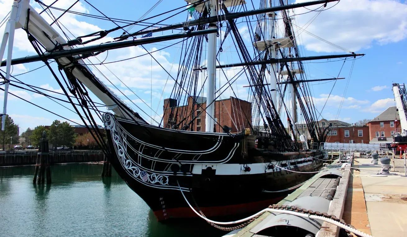 Tall black sailing ship moored in a harbor beside a waterfront promenade and brick buildings