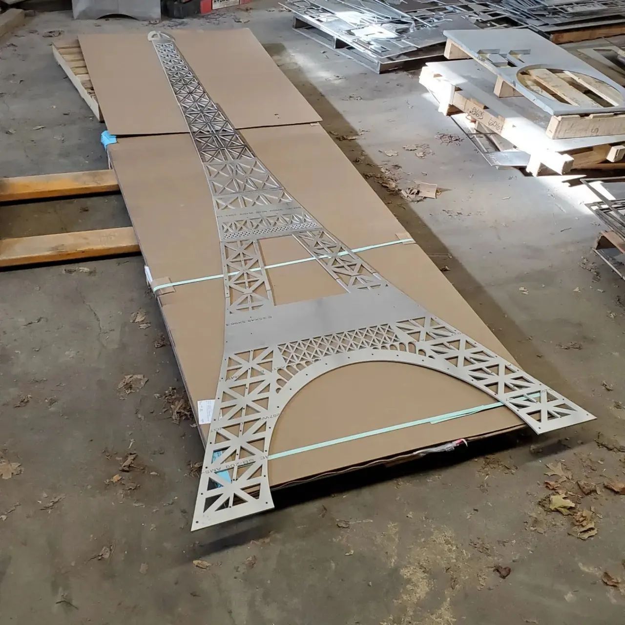 Cardboard template of the Eiffel Tower laid on a workshop floor with white cutout lattice pieces.
