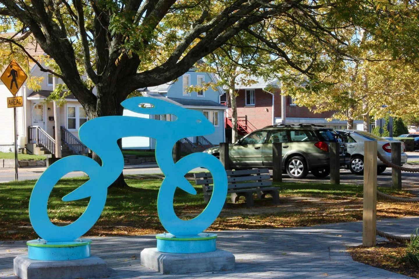 Blue bicycle sculpture on a sidewalk under trees, with parked cars and houses in the background