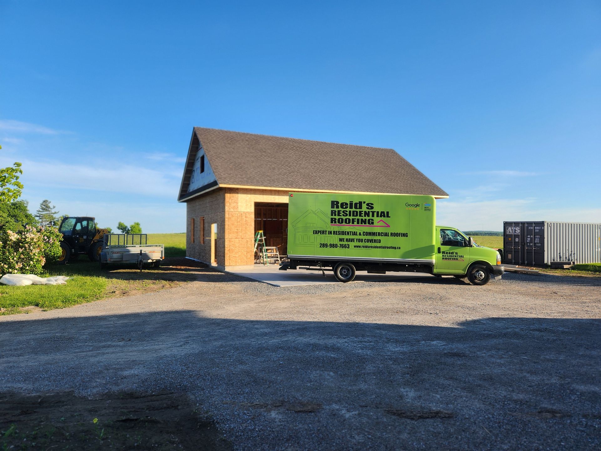 Green moving truck beside a small brick house on a gravel lot under a clear blue sky
