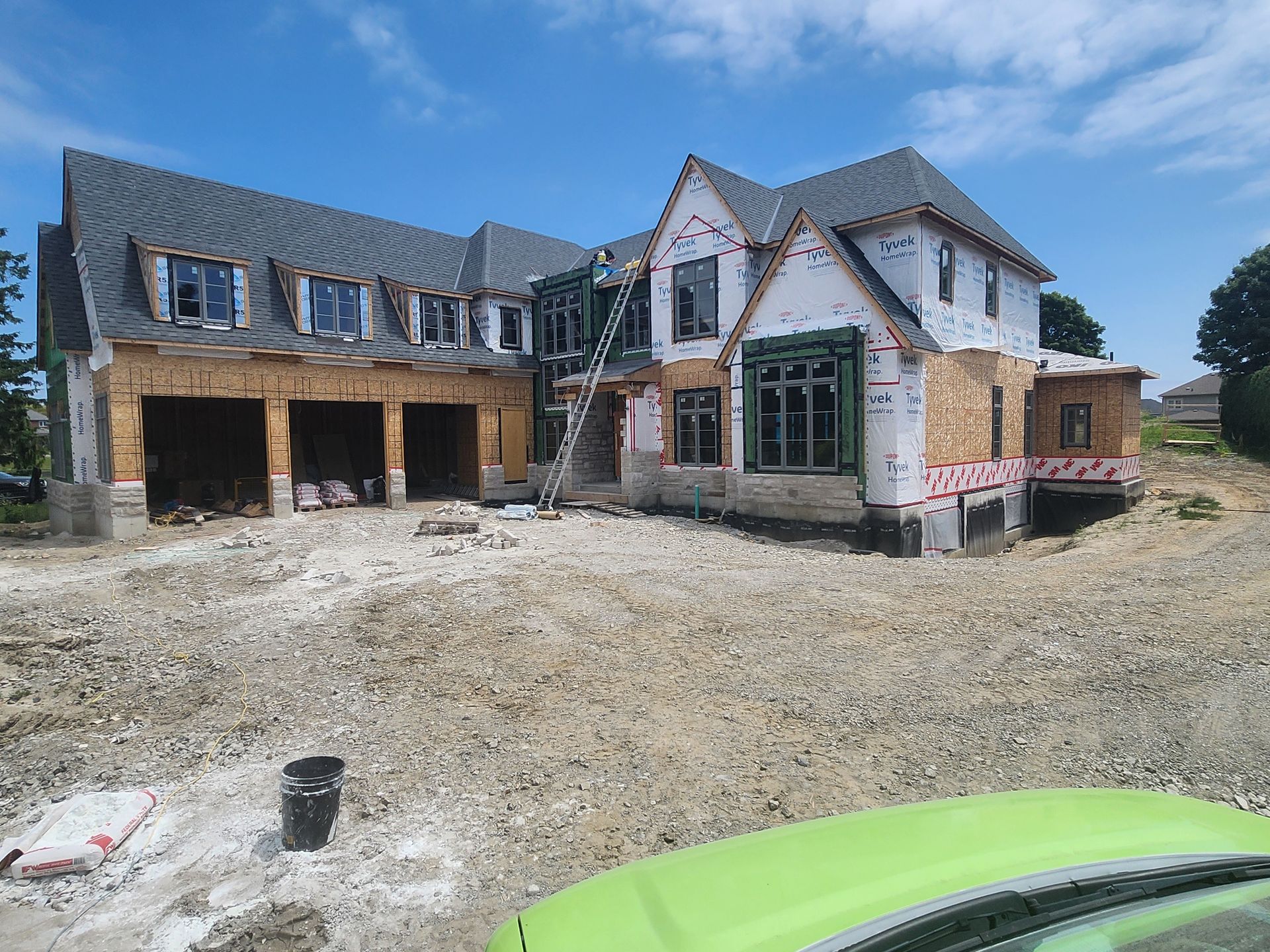 Large unfinished house with stone and wood siding beside a gravel driveway under a clear blue sky