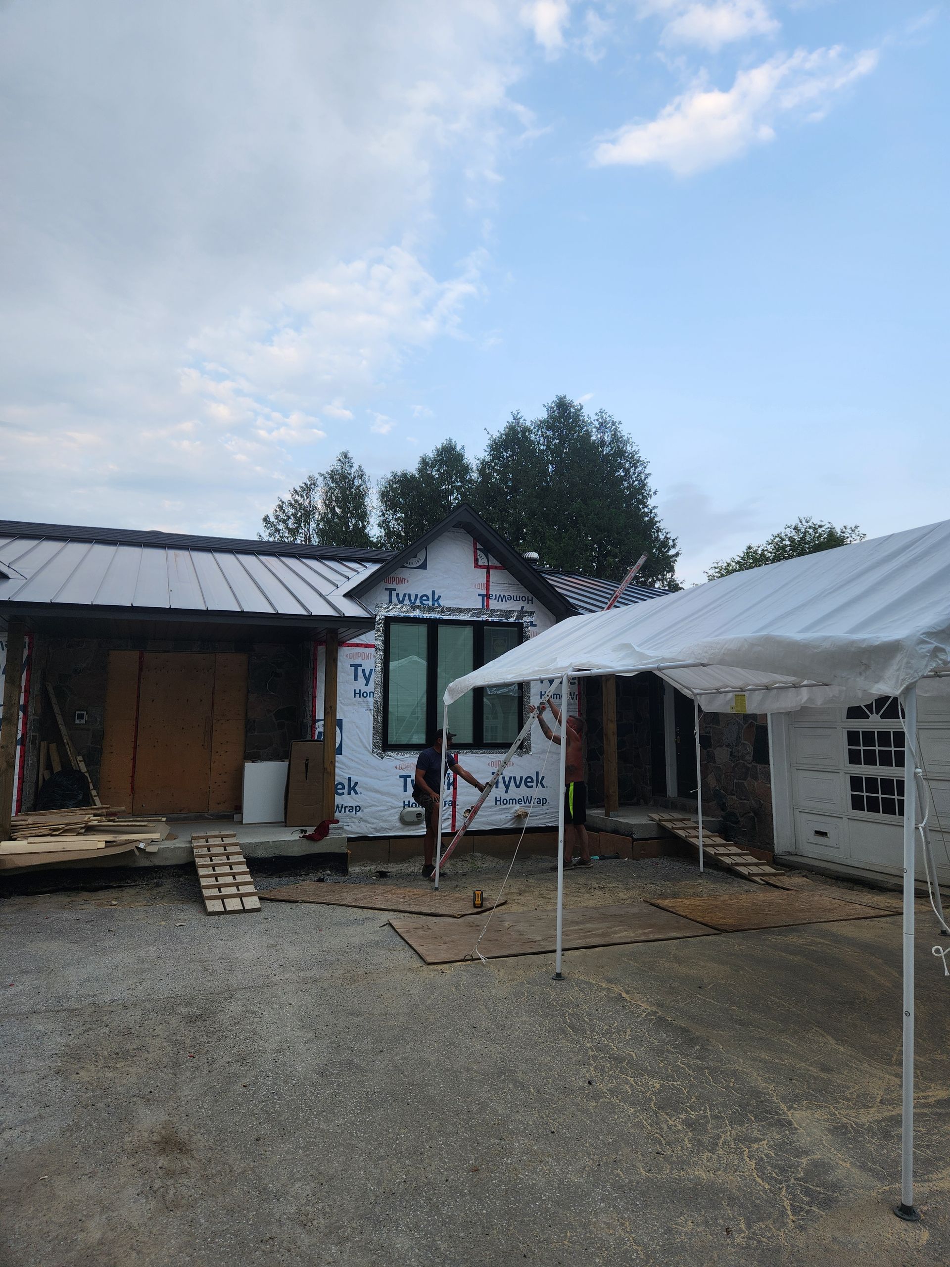 Small rustic buildings with a white canopy in a gravel courtyard under a cloudy sky.