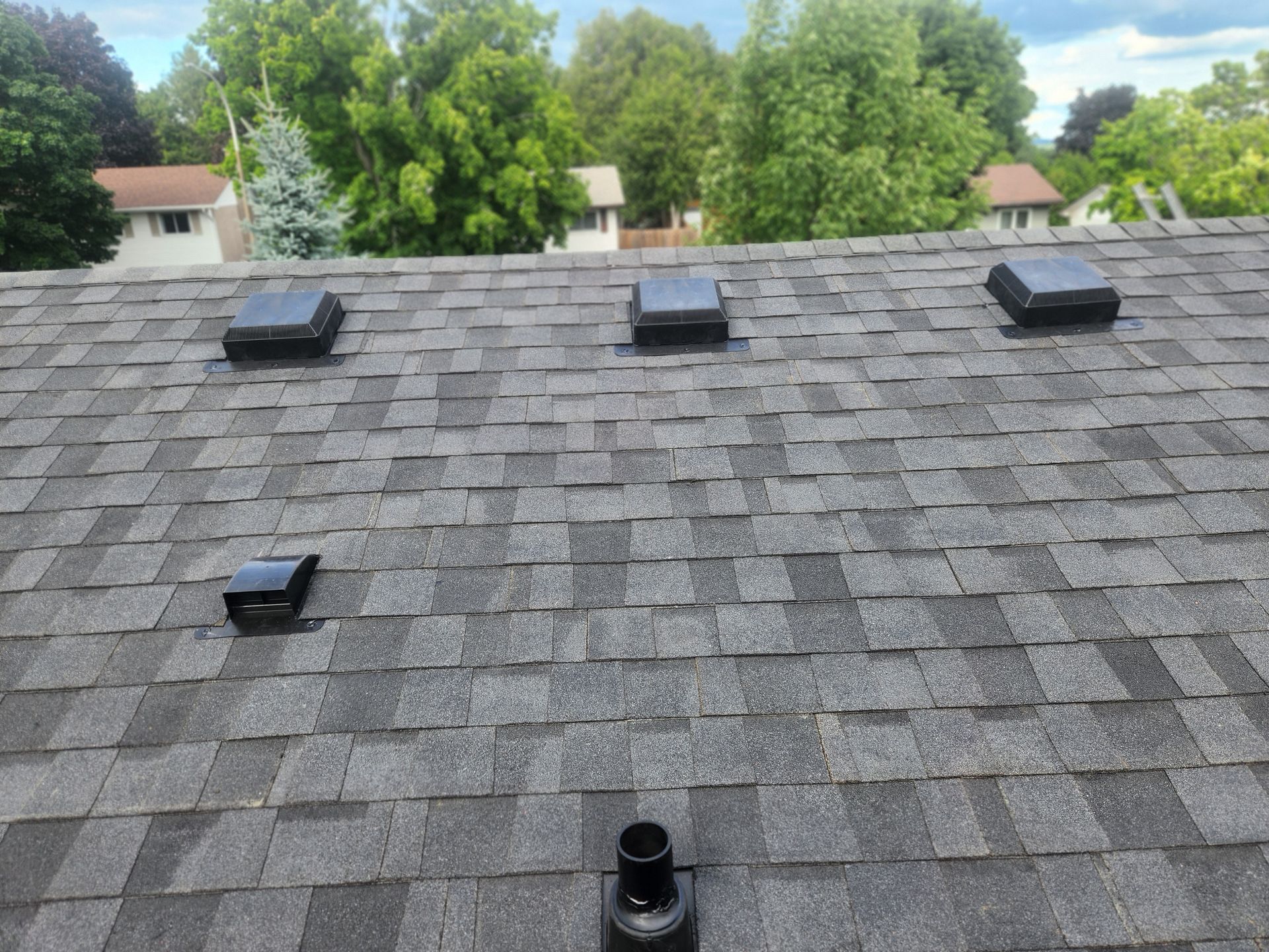 Rooftop with gray shingles, three vents, and trees and houses in the background.