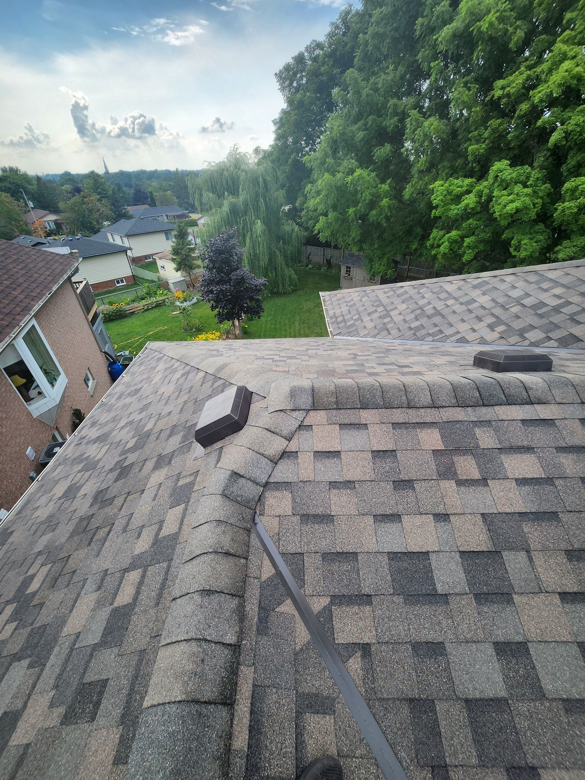 View from a shingled rooftop over a leafy suburban neighborhood under a partly cloudy sky
