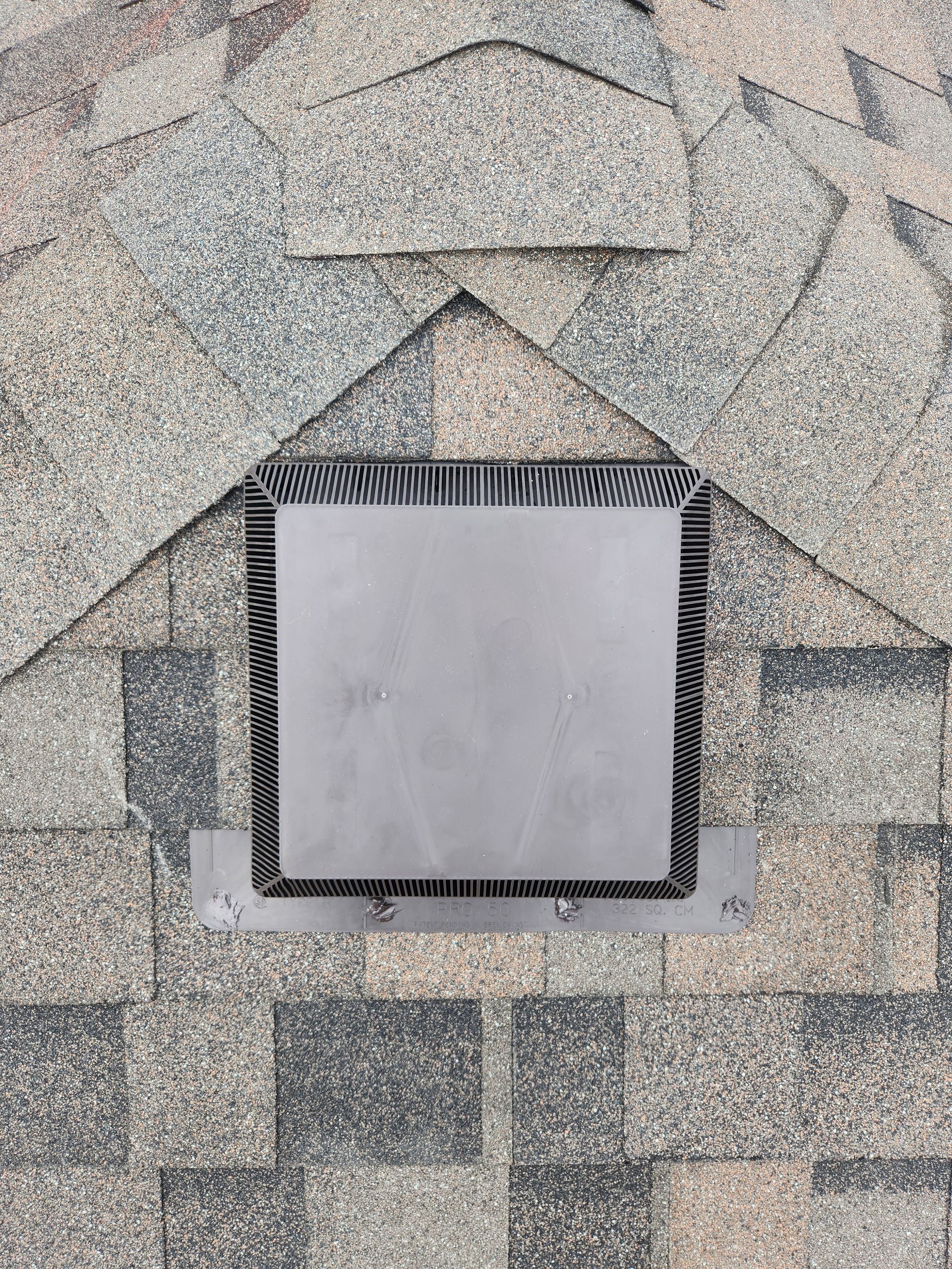 Square gray vent on a shingled roof, centered beneath a triangular roofline.