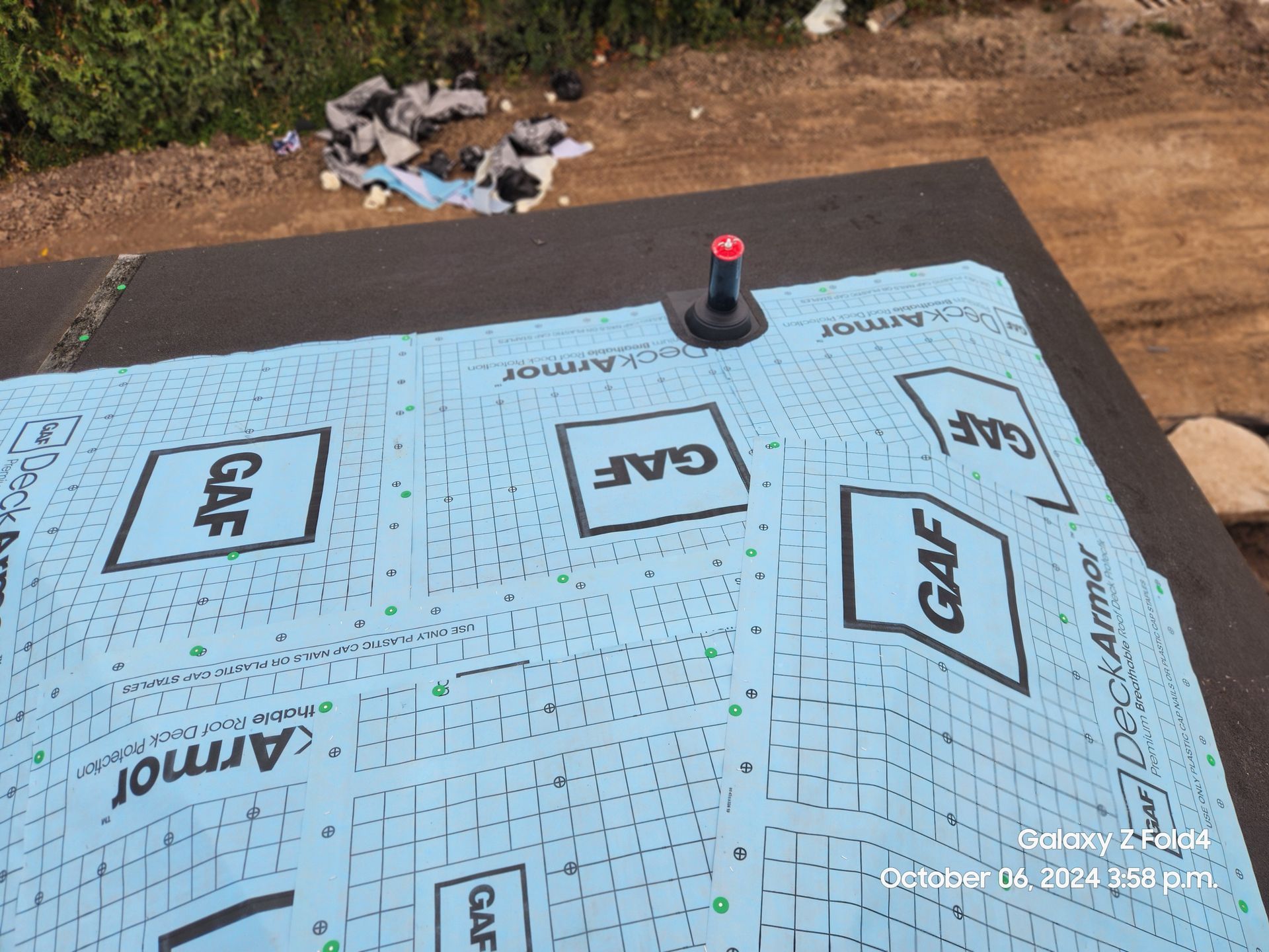 Blue roofing underlayment laid on a roof deck with a small vent pipe and a dirt yard in the background