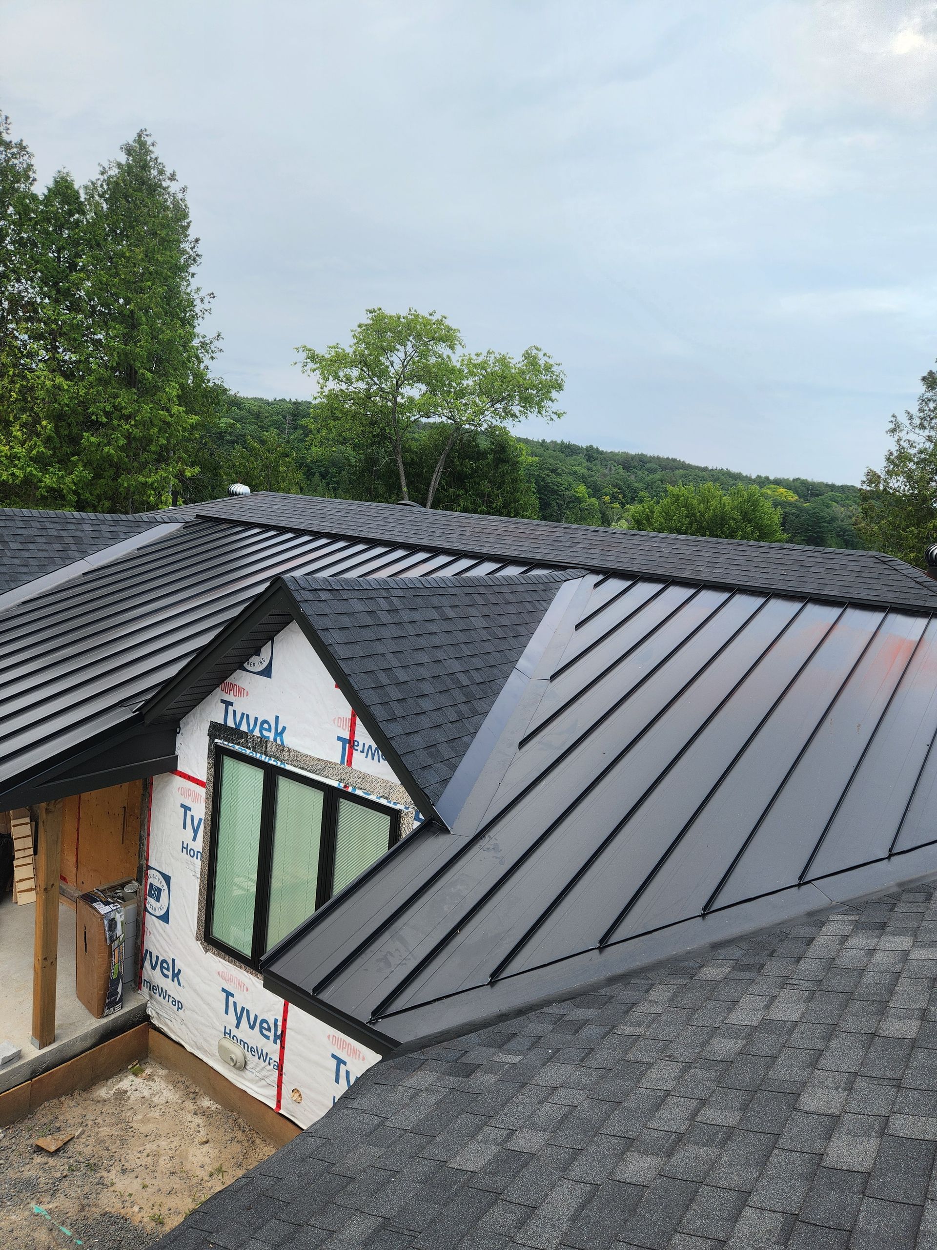 New dark metal roof on a house under construction, with visible roof windows and surrounding trees.