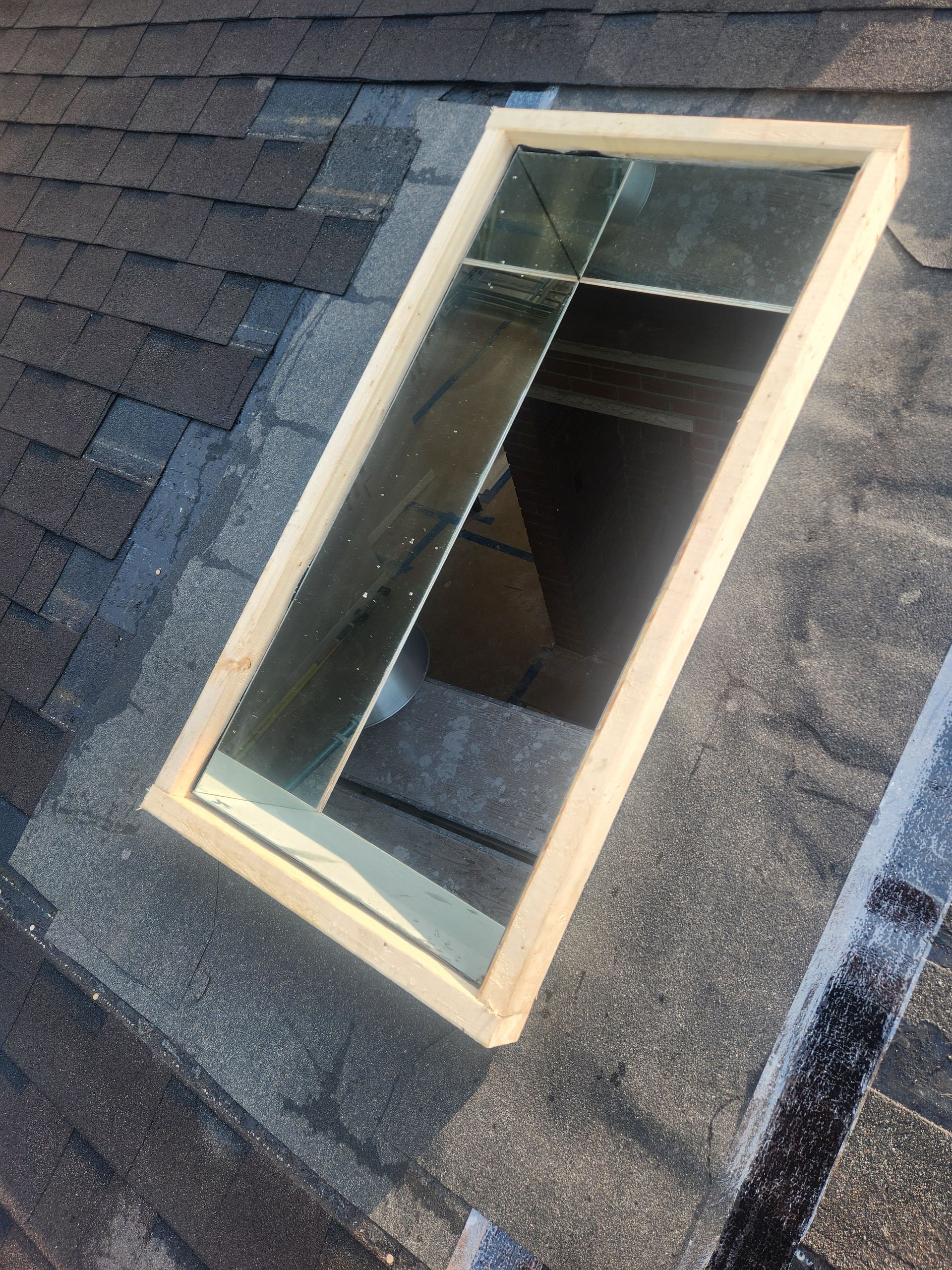 Skylight window with a wooden frame on a shingled roof, reflecting light.