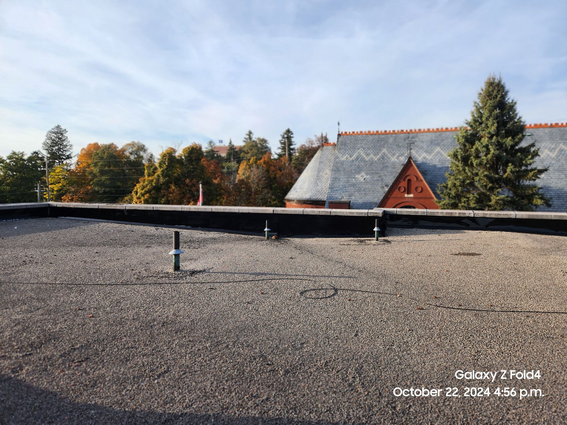 Flat rooftop with gravel, skylight vent, and a red house and trees beyond a low wall at sunset.
