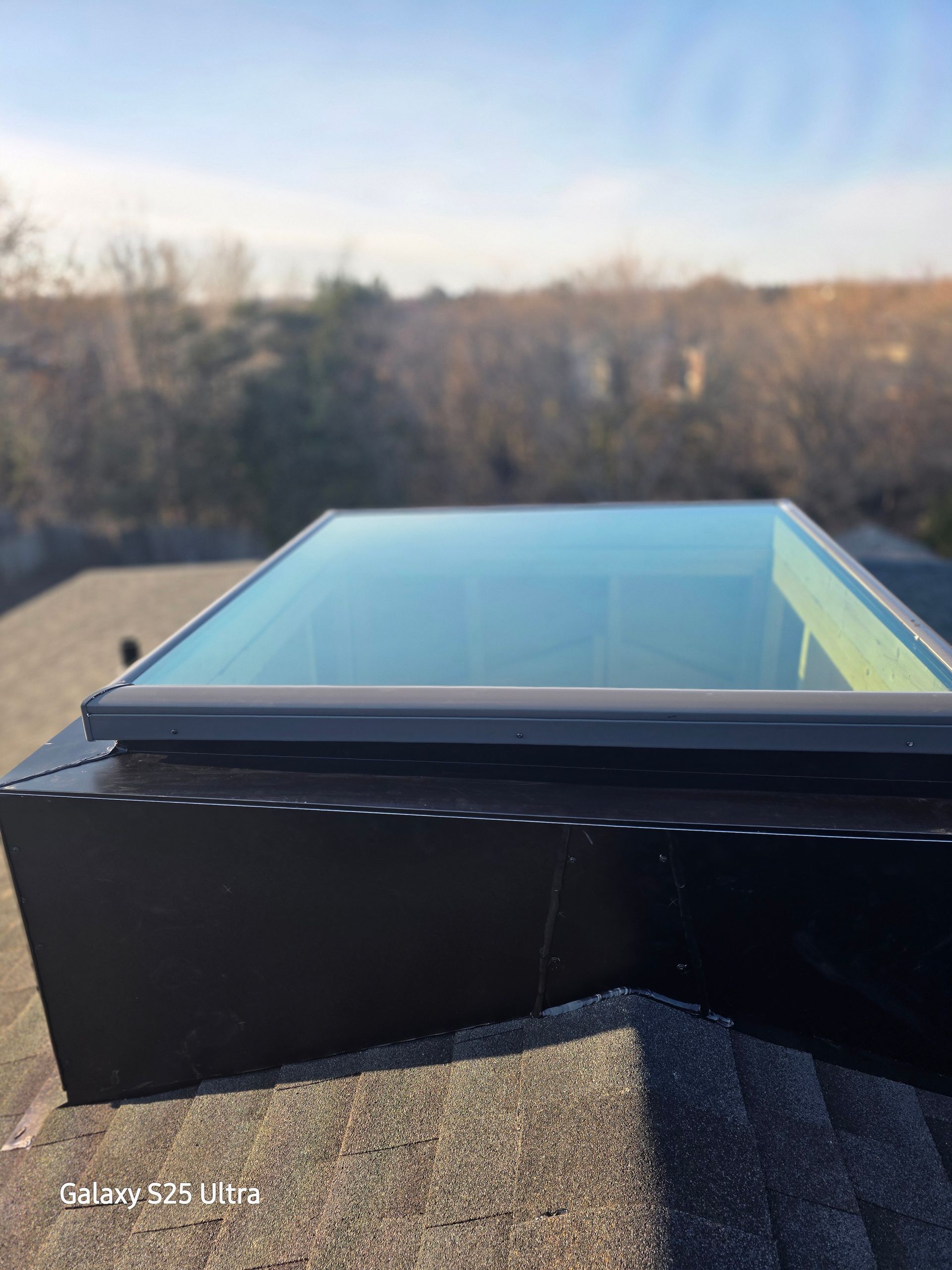 Black rooftop skylight with glass panel against a blurred outdoor background at sunset