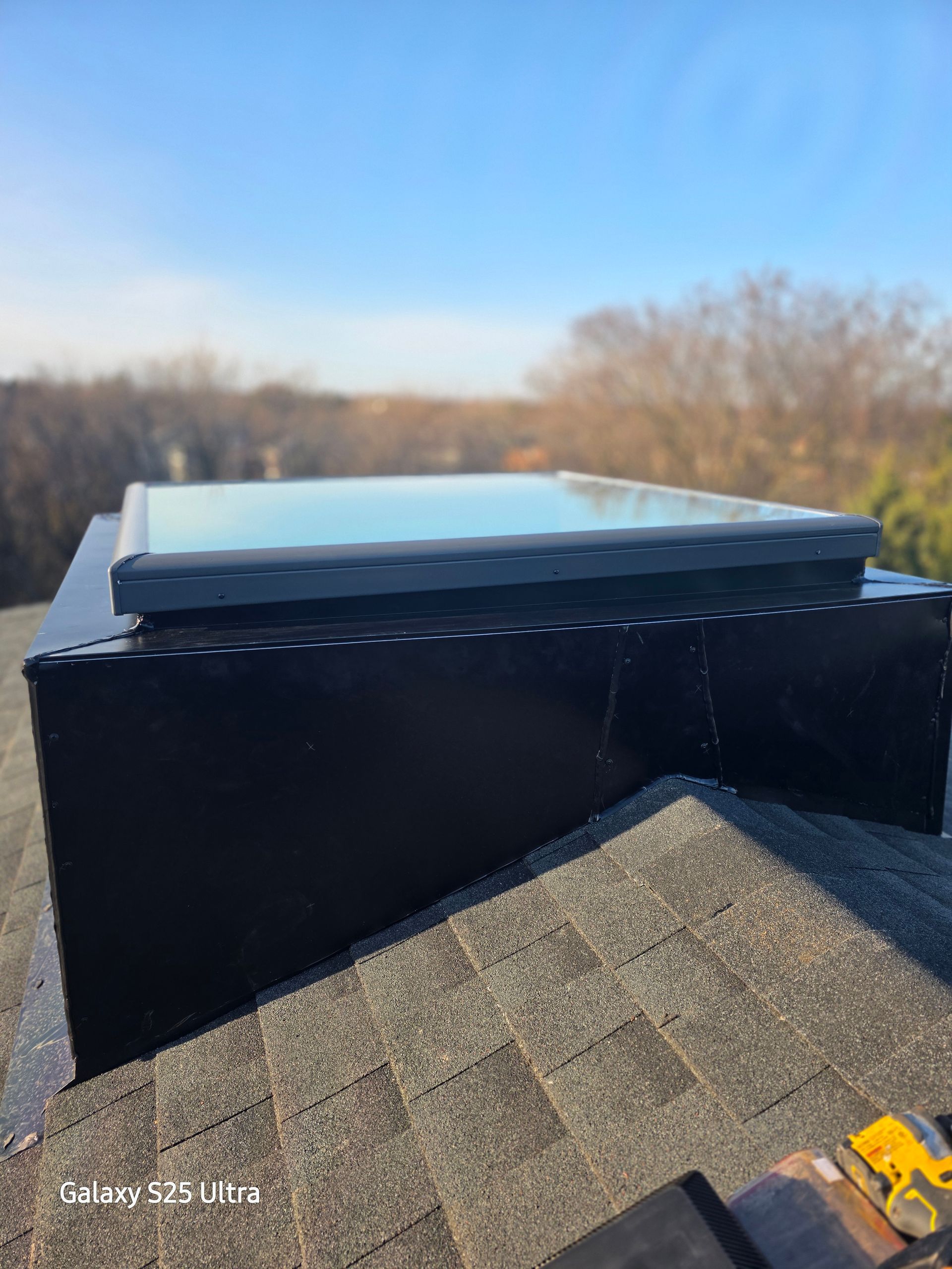 Black rooftop with a skylight under a clear blue sky, with trees in the background.
