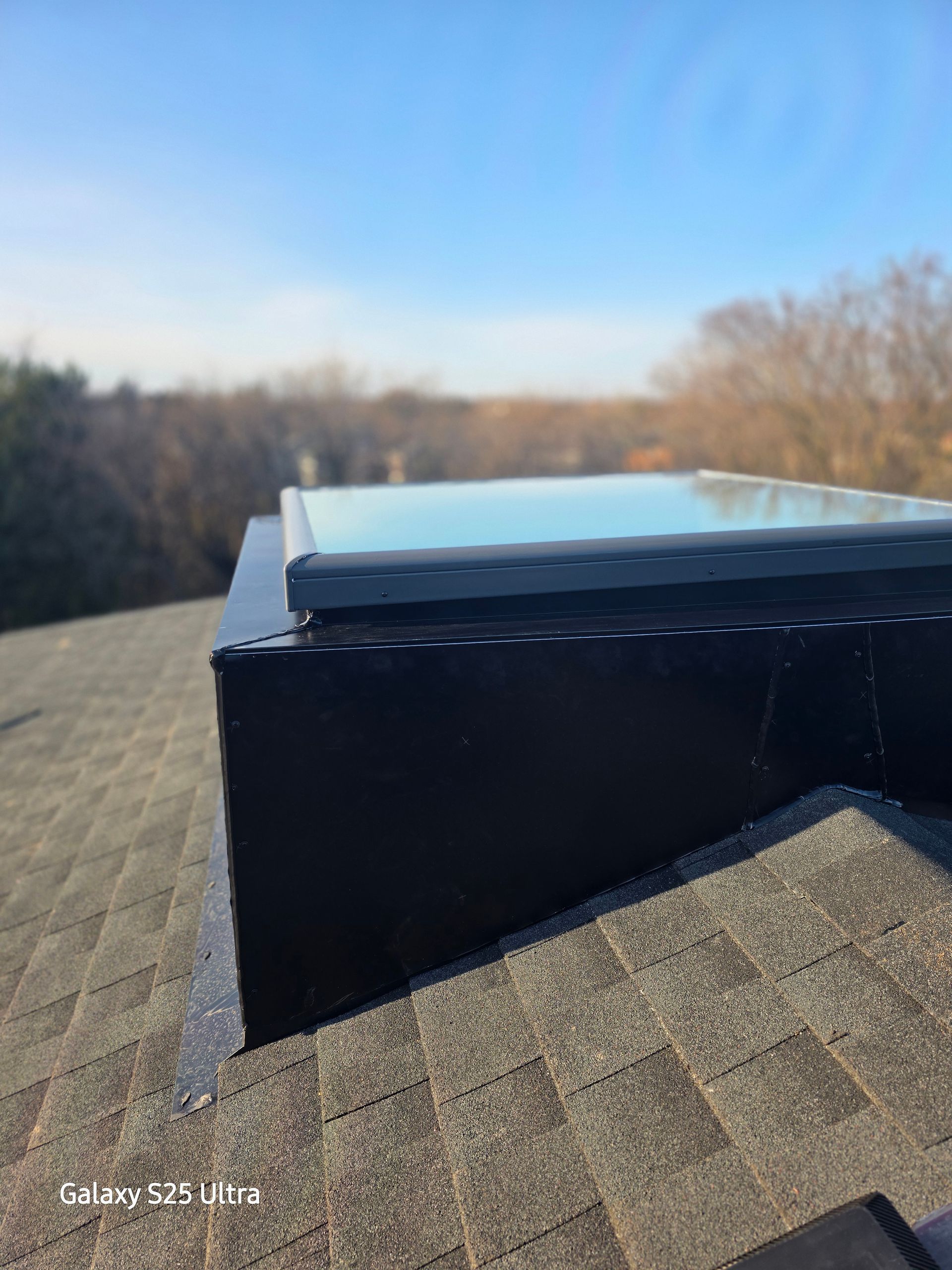 Black roof vent or skylight on a shingled roof under a clear blue sky