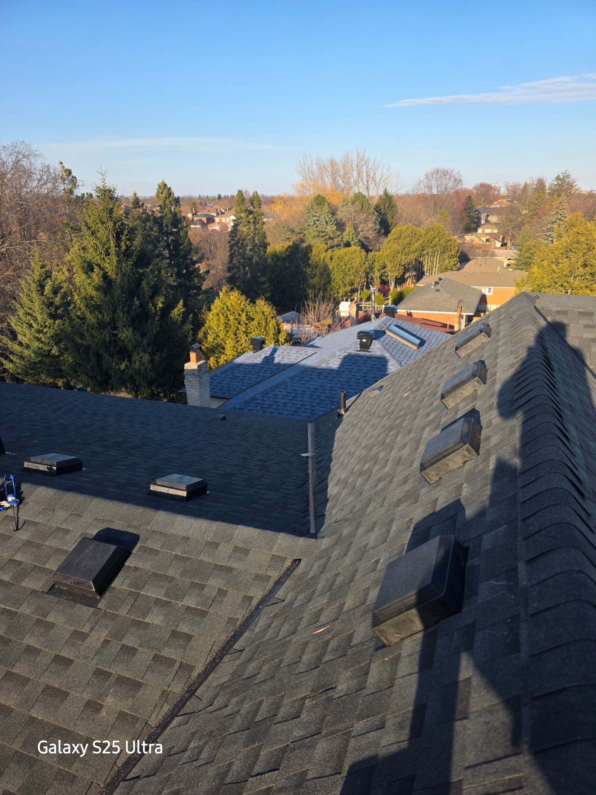Rooftop view of suburban houses and trees under a clear sky, with shingles in the foreground.