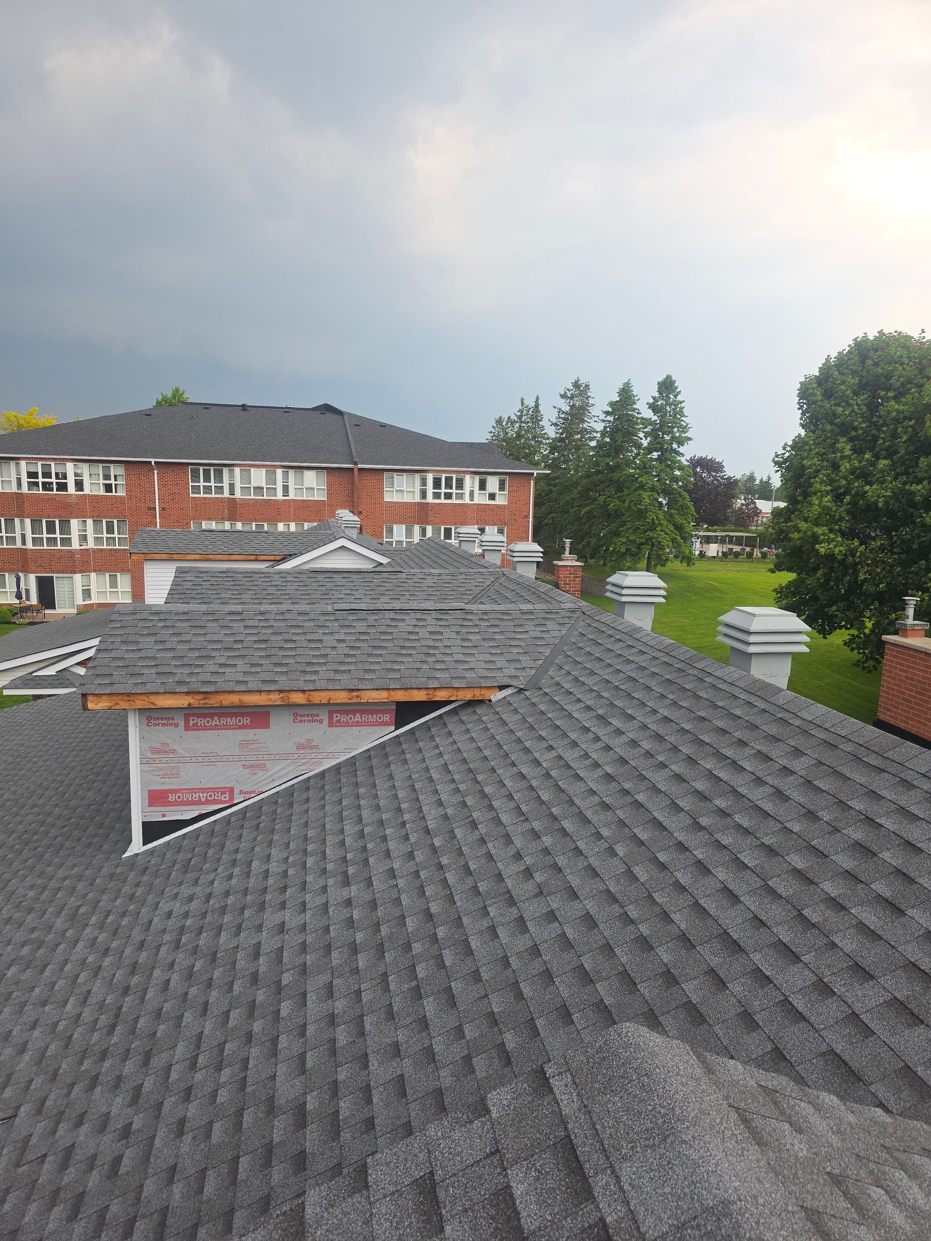 Rooftops with dark gray shingles overlooking red-brick buildings, trees, and a cloudy sky