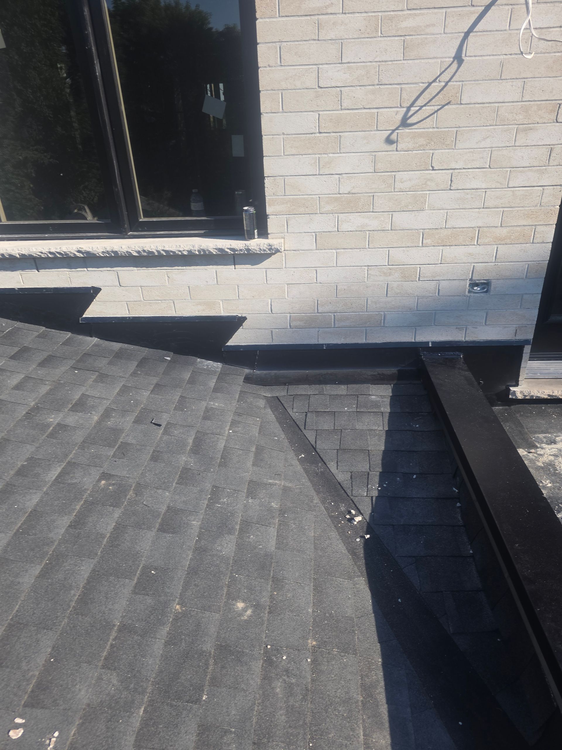 Rooftop with dark shingles and a brick wall, showing a damaged or uneven edge near a corner.