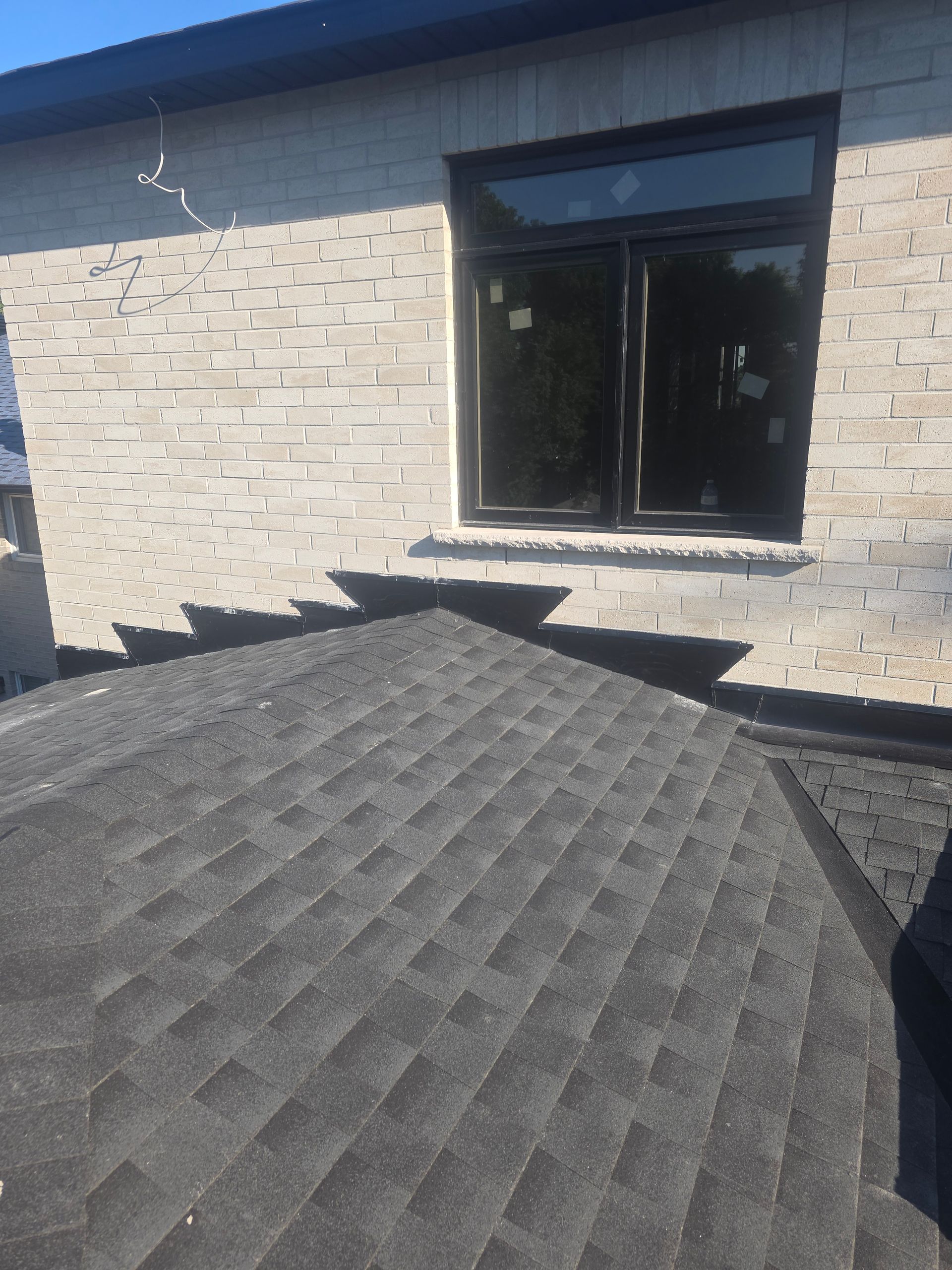 Black shingled roof section against a brick house with a dark-framed window in sunlight
