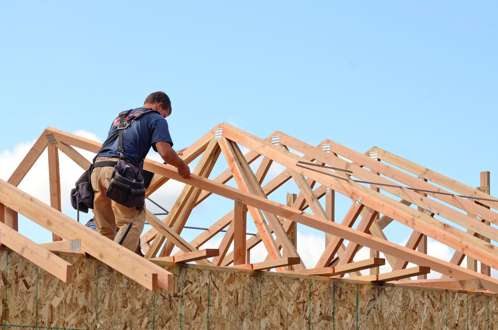 Worker on a roof frame installing wooden trusses under a clear blue sky.