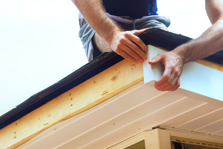 Hands installing a white soffit under a roof eave with a wooden beam.