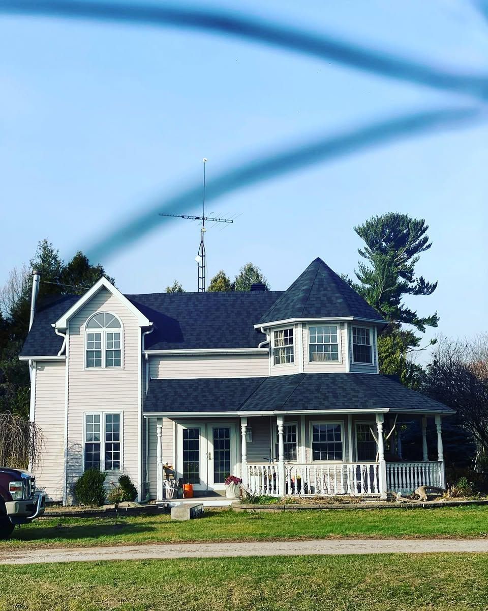 Two-story white house with dark roof and wraparound porch on a grassy lot under a clear blue sky