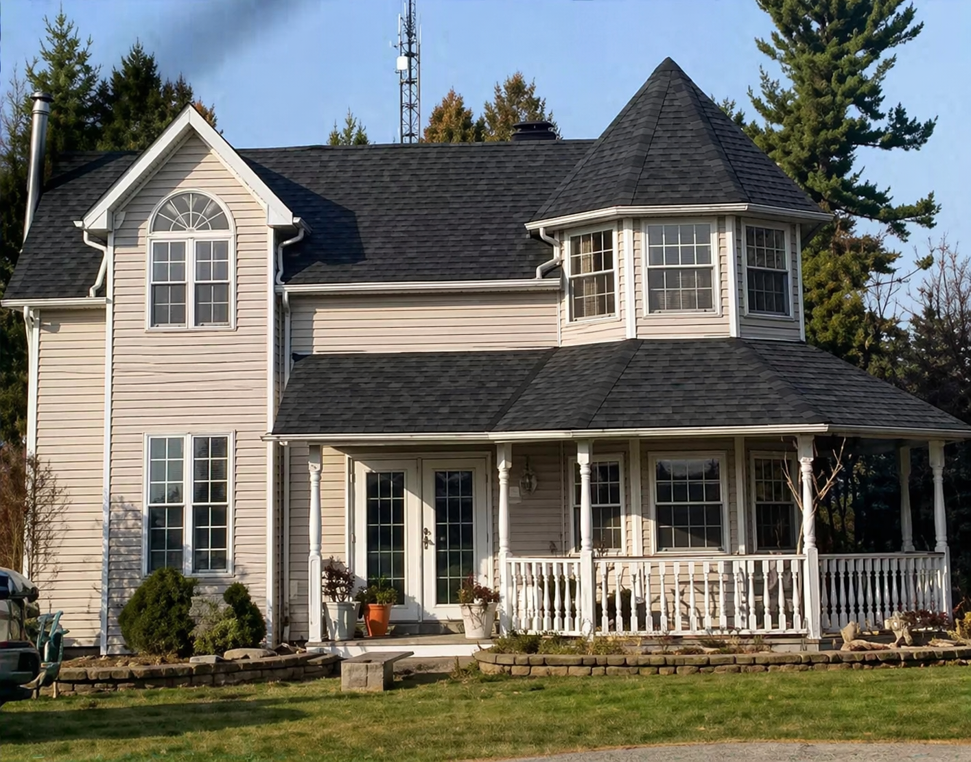 Two-story beige house with dark shingle roof and white porch railings, surrounded by trees and lawn