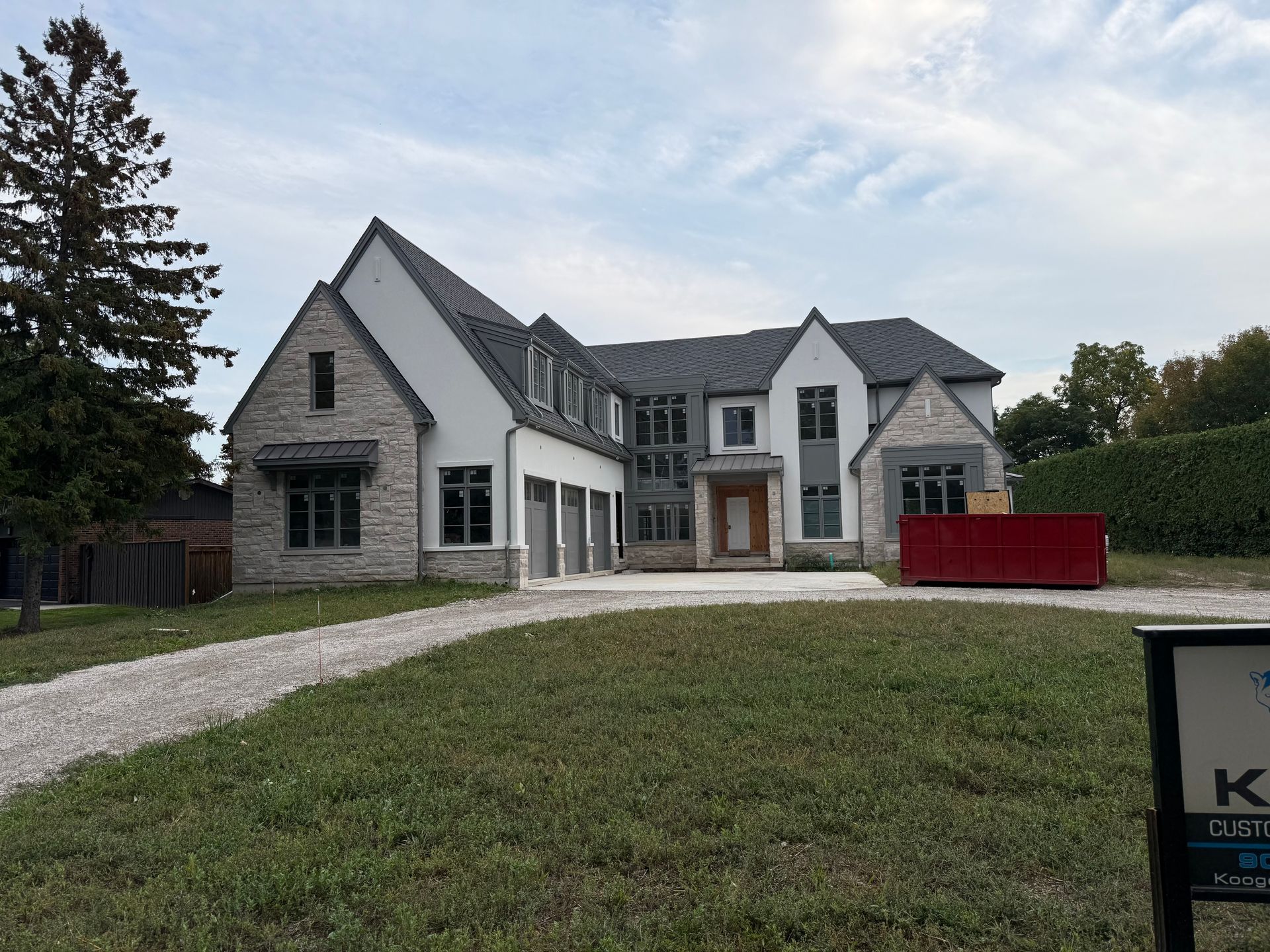 Large gray house with a gravel driveway, red dumpster, and a grassy front yard under a cloudy sky