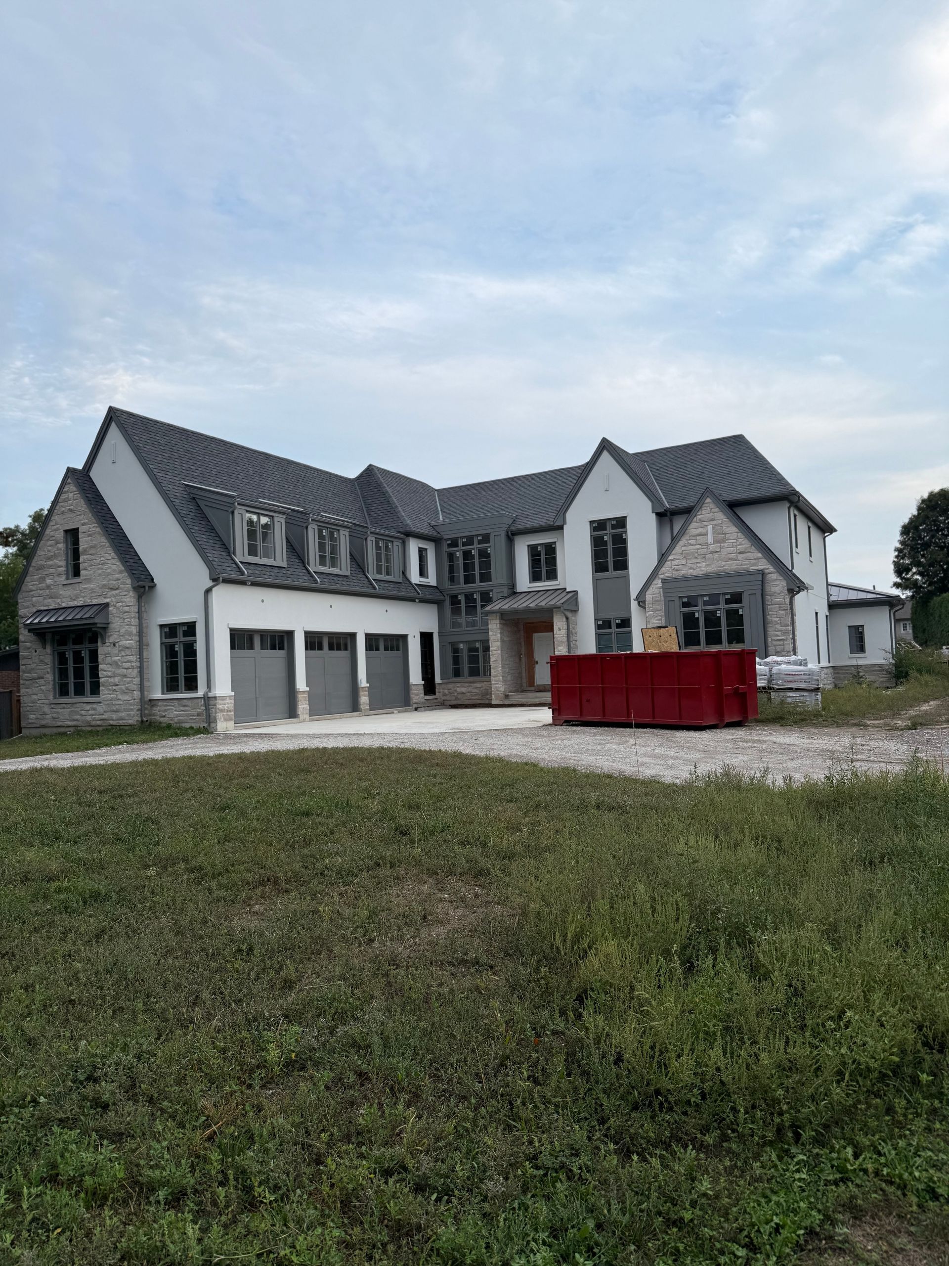 Modern gray house with multiple gables, garage doors, and a red dumpster in the driveway under a cloudy sky