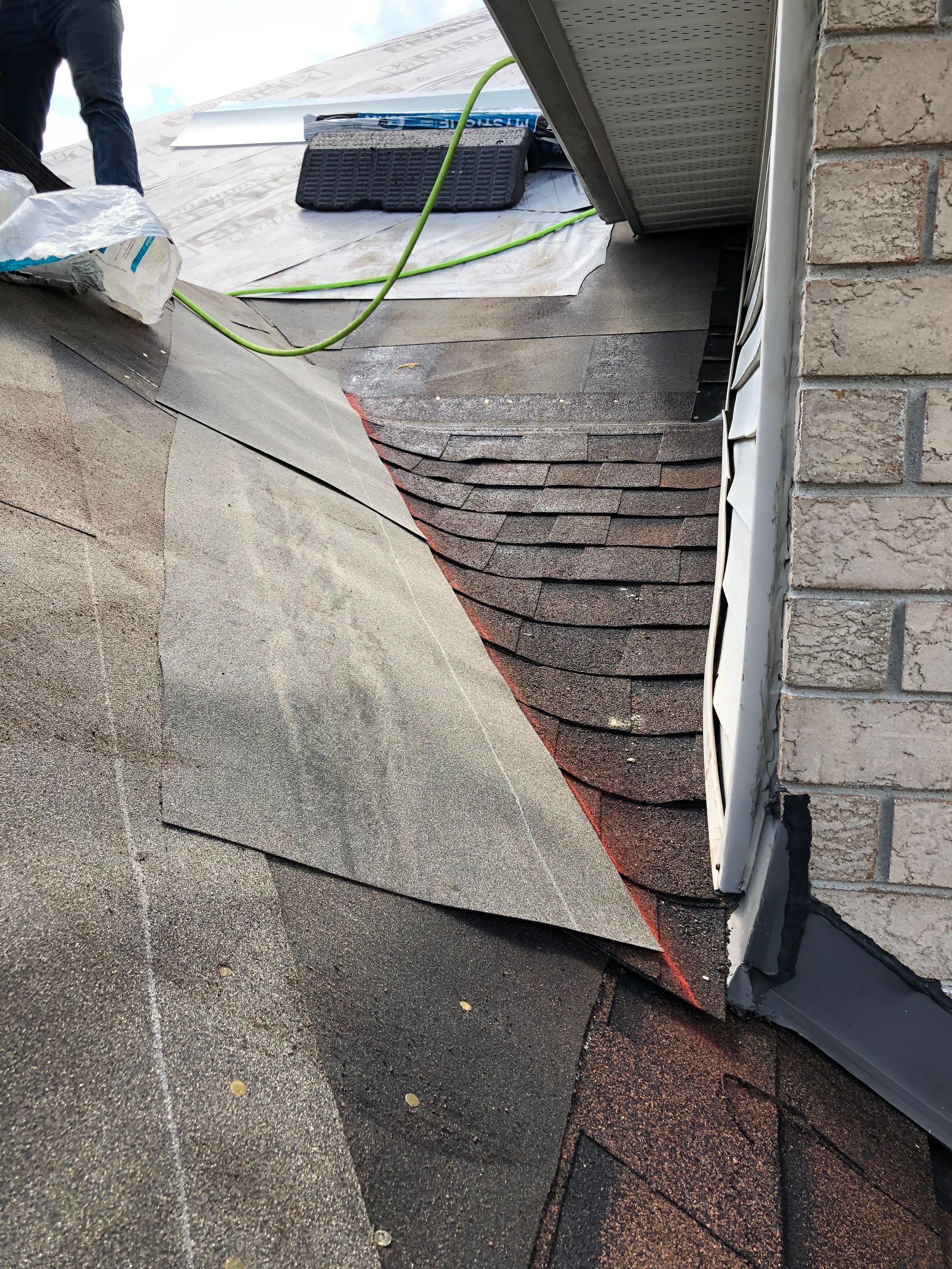 Damaged roof shingles at a steep valley near a chimney and siding, with exposed layers and debris