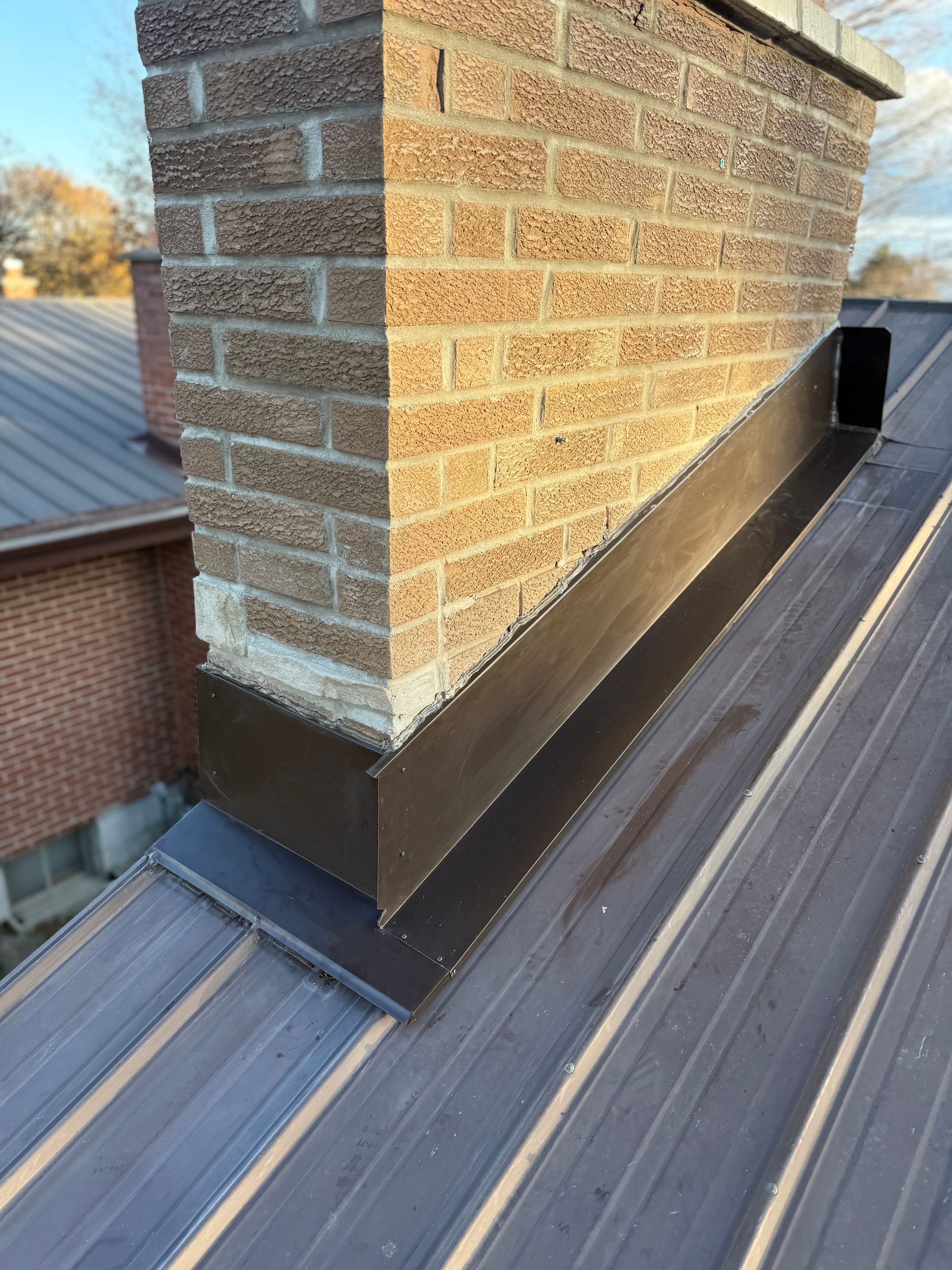 Brick chimney on a dark metal roof with a flashing base and rooftop vent nearby