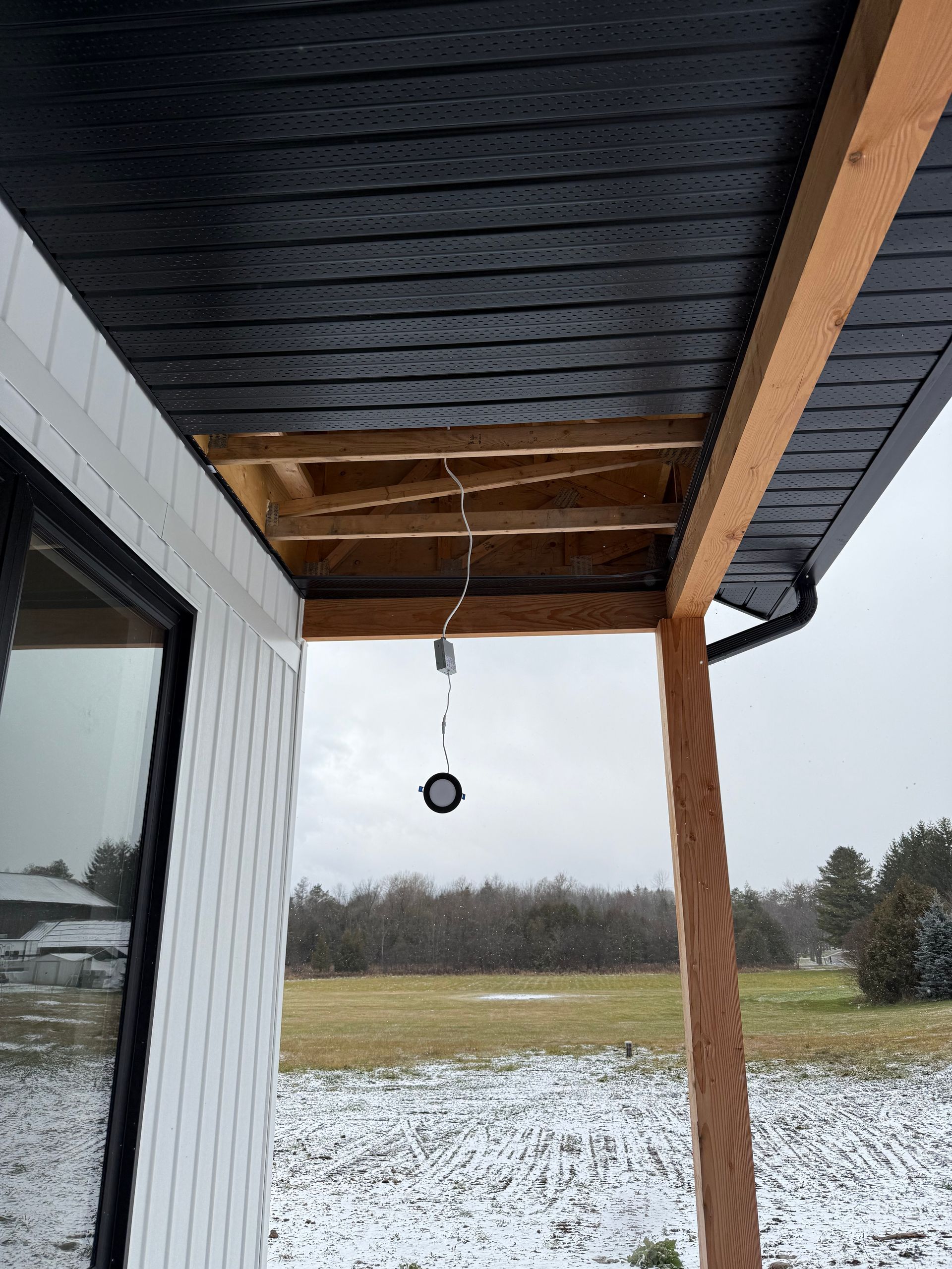 Snowy backyard view from a porch, with a hanging black ornament under a dark roof and wooden support post.