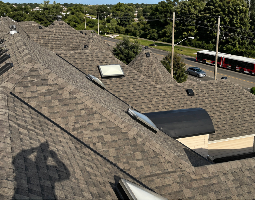Rooftops with gray shingles and skylights beside a road with cars and trees