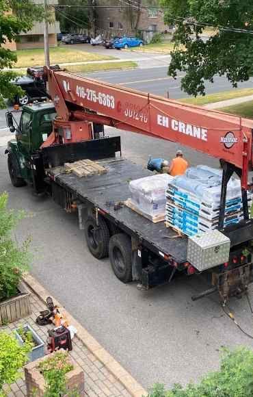 Crane truck lifting stacked materials on a street beside a parked flatbed truck