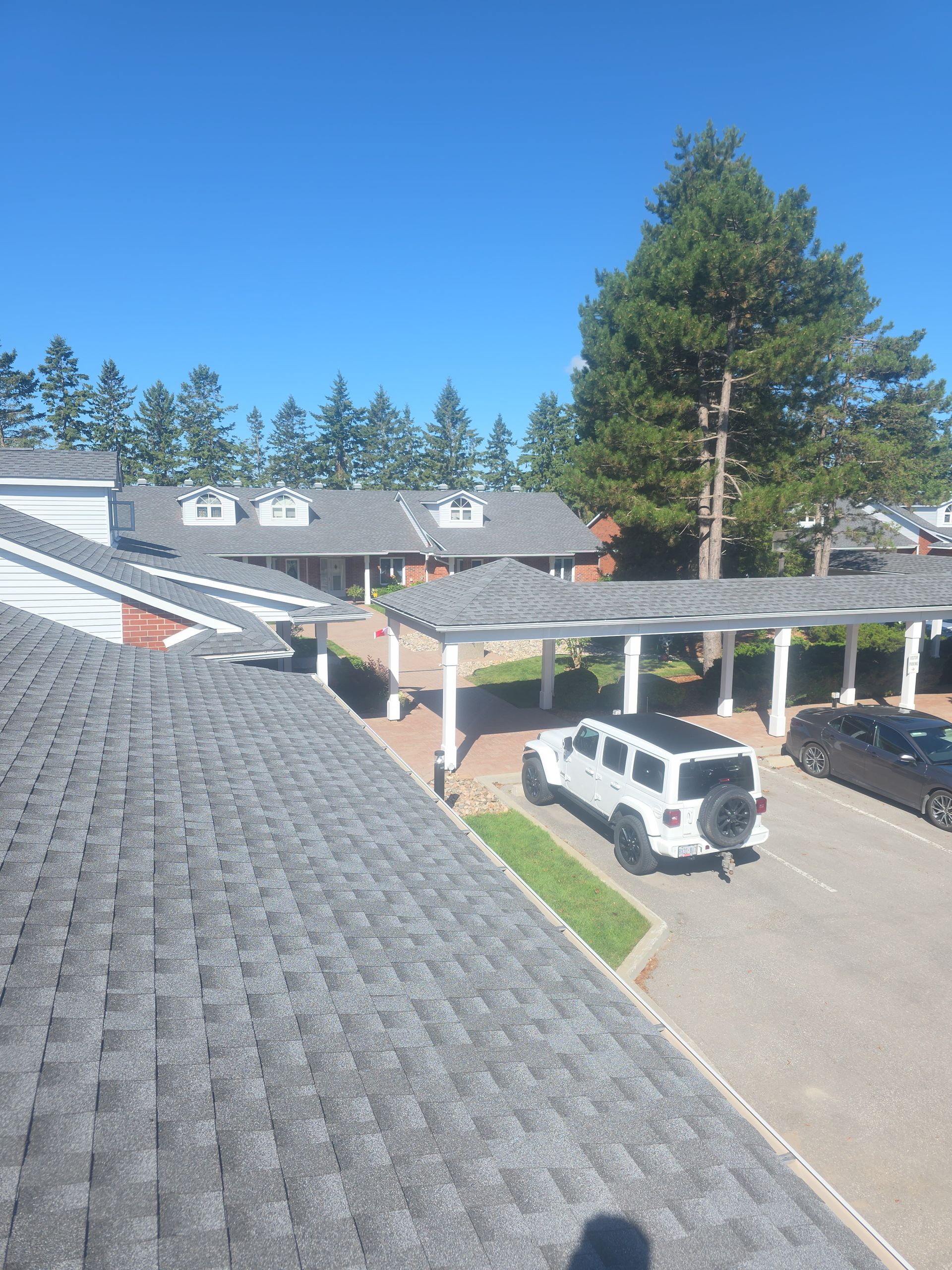 Apartment complex parking lot with white SUV, covered carport, gray roof, and trees under a blue sky