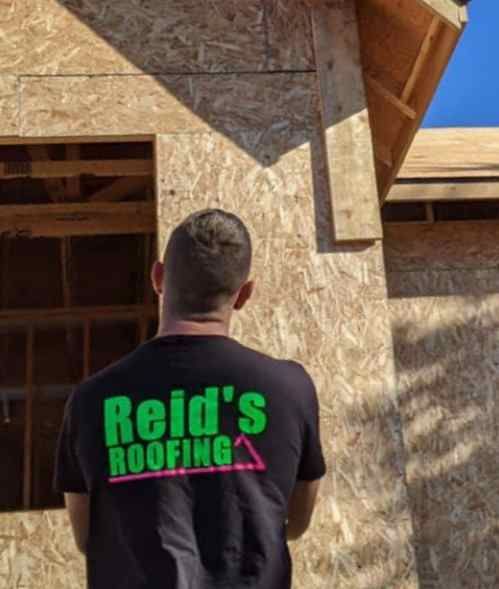 Person in black shirt with neon green “Reid’s Roofing” logo stands beside a house under construction.