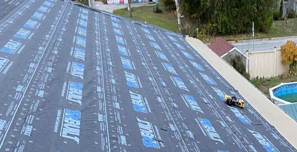 Worker on a steep roof installing black roofing felt near a driveway and landscaping.