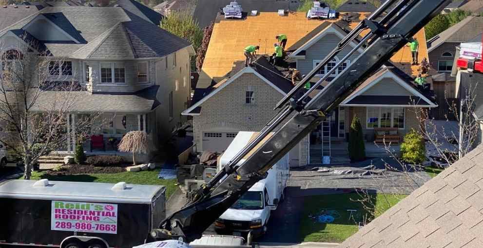 Large black crane extends over parked white van in a suburban neighborhood with houses and a moving truck.