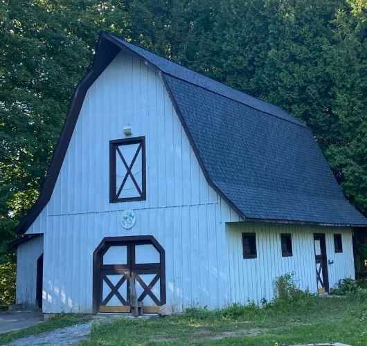 White barn with a steep dark roof and X-braced doors, set against trees in a grassy area.