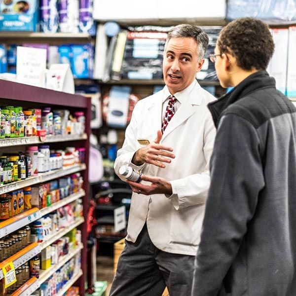 Pharmacist talking with a customer in a pharmacy aisle, holding a bottle and gesturing toward stocked shelves