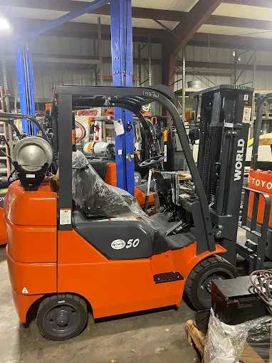 Orange forklift in a warehouse with a driver inside, parked near another forklift.