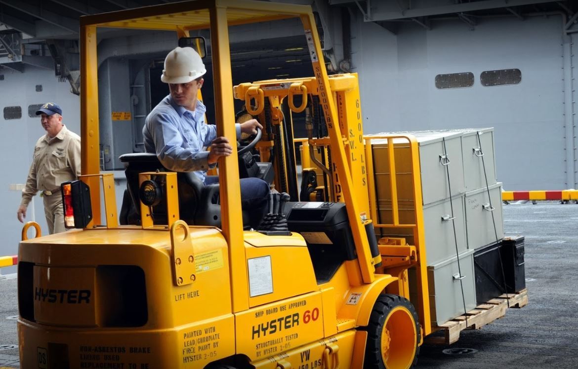 Yellow Hyster forklift moving crates in an industrial warehouse, with a worker standing nearby