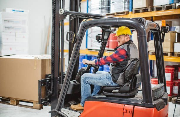 Forklift operator in a warehouse moving a pallet beside stacked boxes