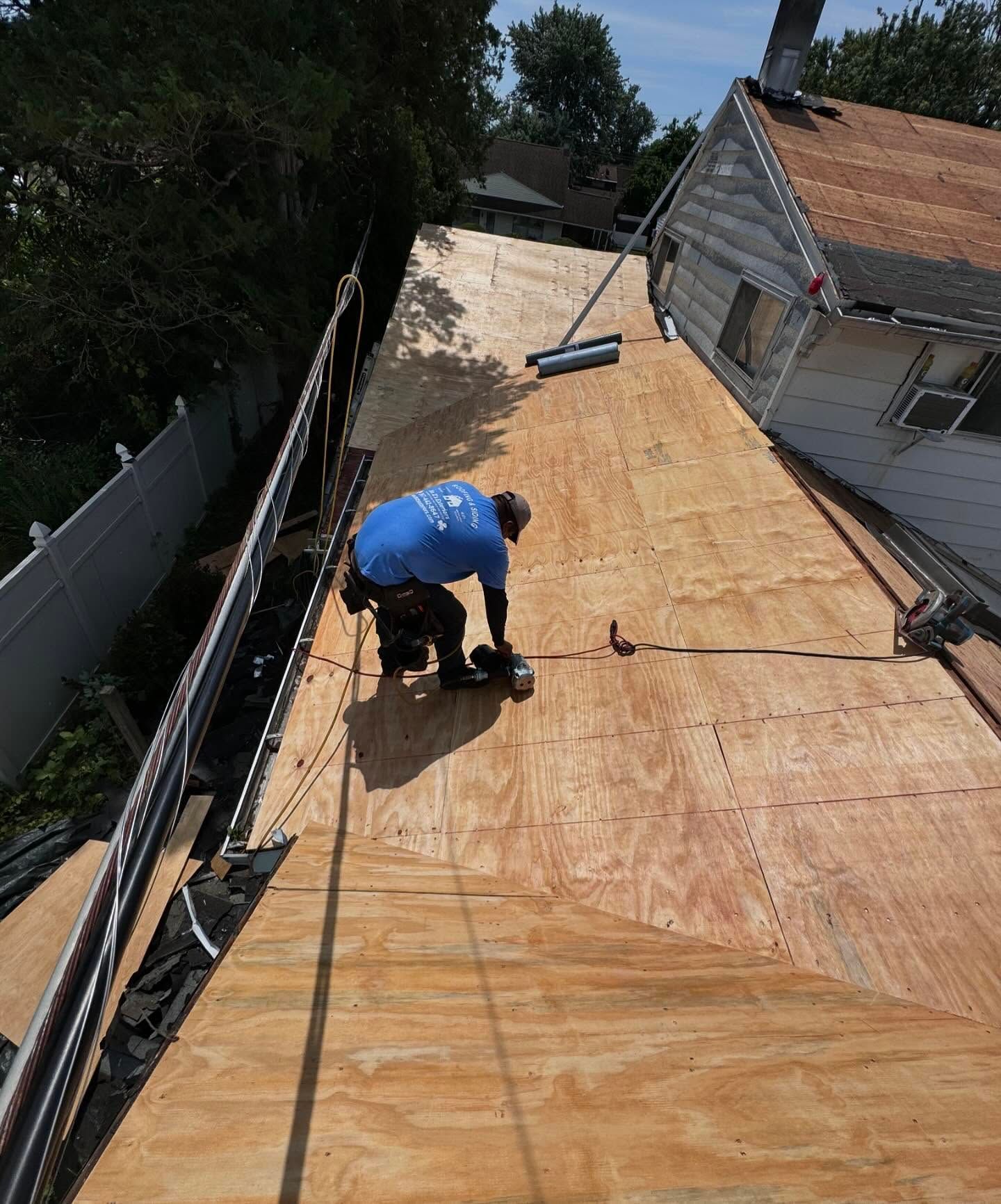 Worker in blue shirt nailing plywood sheathing on a roof beside a house.