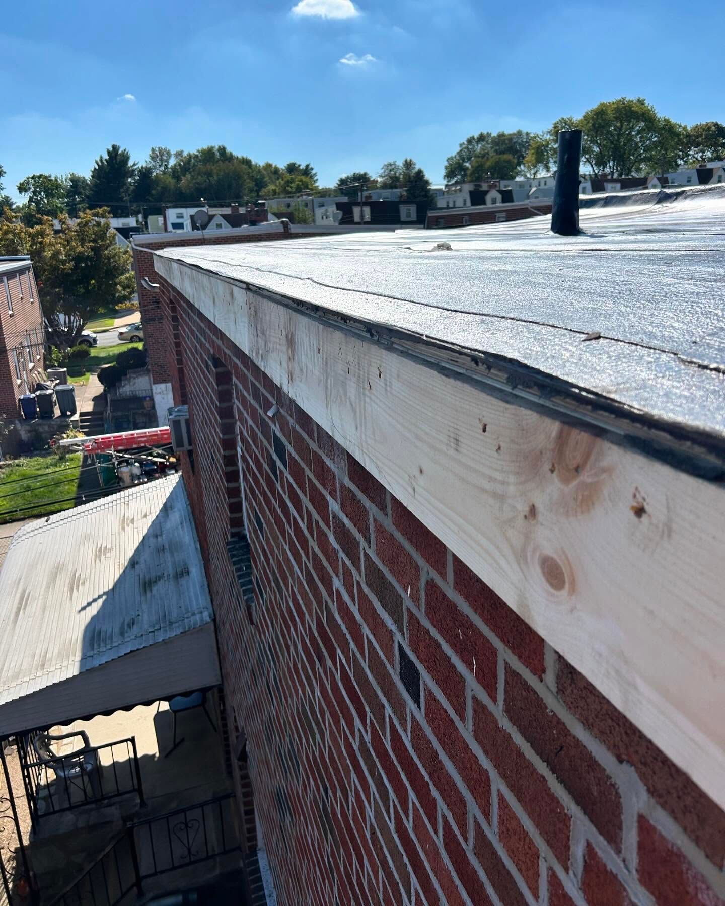 Brick building rooftop with tar paper, wooden trim, and a vent pipe under a blue sky