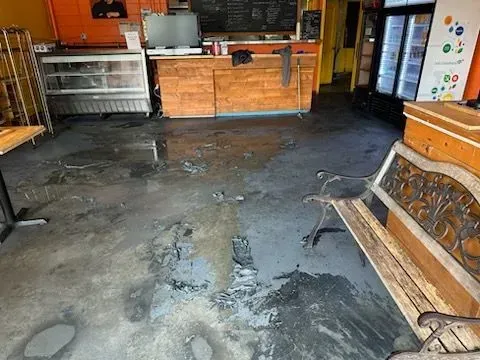 Interior of a business with a wet, damaged concrete floor; counter and refrigerator are visible.