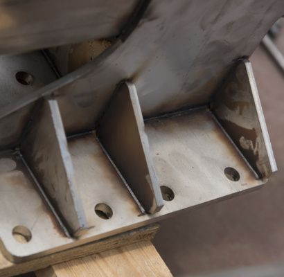 Primo piano di una staffa metallica saldata con rinforzi triangolari e fori di montaggio su una superficie di legno.