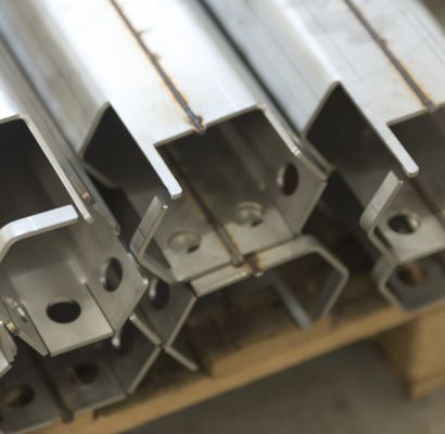 Travi metalliche argentate impilate, con fori per bulloni, disposte su uno scaffale o un pallet.
