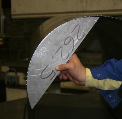 Una mano che regge una lastra di metallo argentato curva, contrassegnata da numeri gessati, in un'officina.
