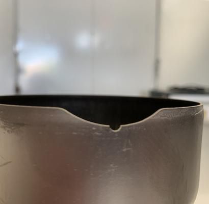 Contenitore in metallo nero con una tacca sul bordo, mostrato in primo piano su uno sfondo interno sfocato.