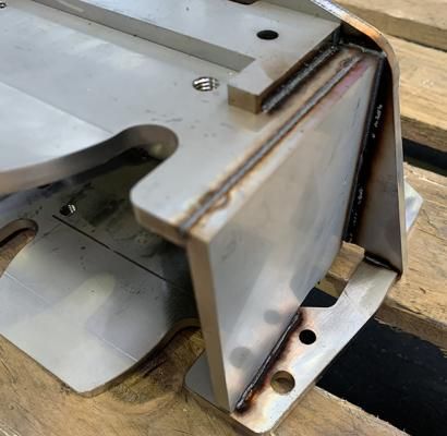 Primo piano di un gruppo di staffe metalliche saldate su un banco da lavoro in legno, che mostra una piastra d'acciaio piegata e gli elementi di fissaggio.