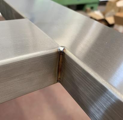 Primo piano di un giunto angolare in metallo spazzolato con saldatura visibile e leggera decolorazione dovuta alla ruggine.